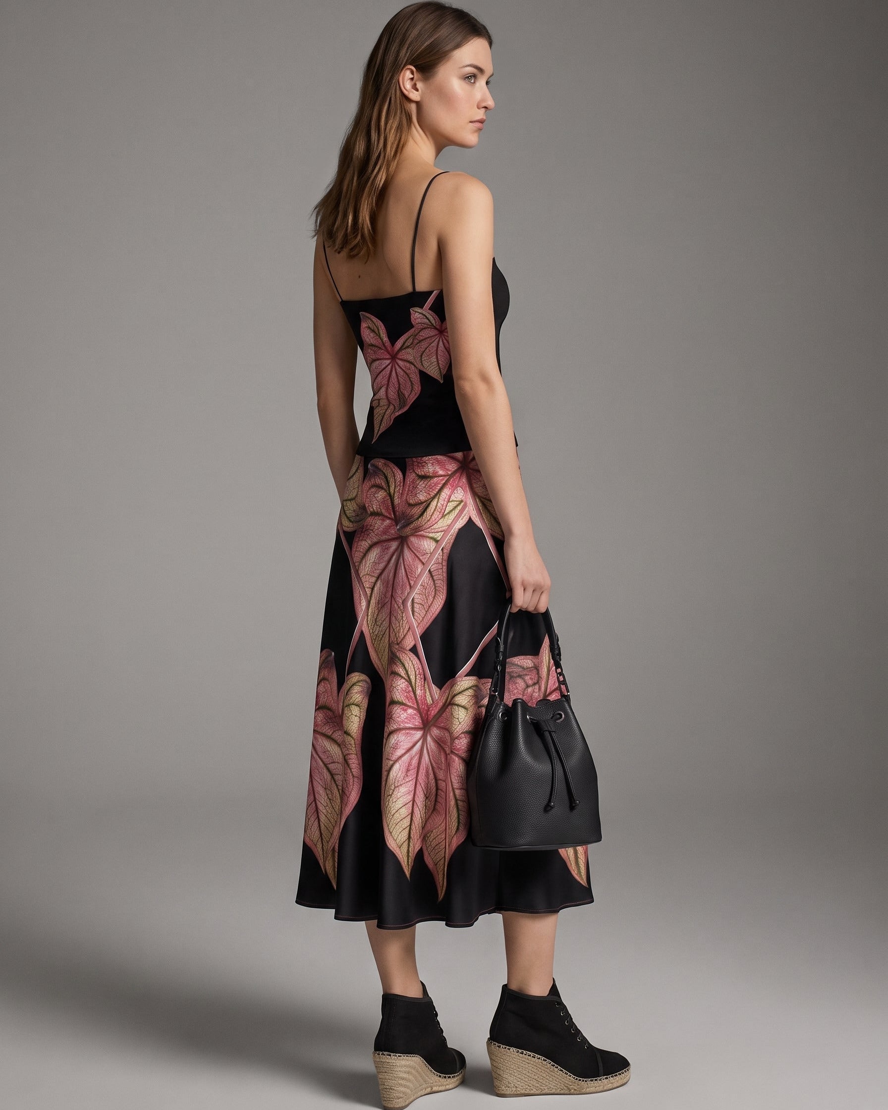 A woman modeling a black midi dress with a pink and gold tropical leaf print, shown from a side-back angle against a neutral gray studio background.