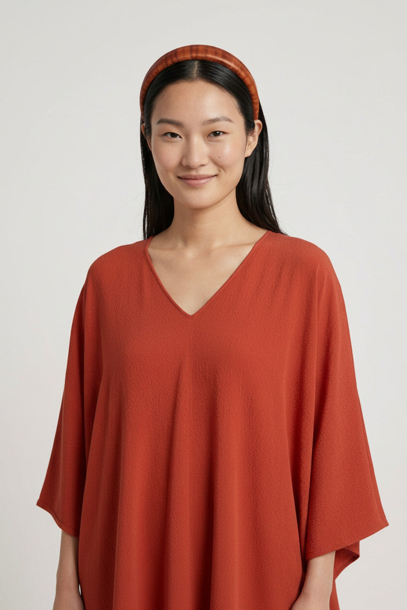 A woman wearing a rust-orange V-neck top and a matching headband, shown from the back of her head to the waist against a light gray studio background. Another person is partially visible in the foreground.